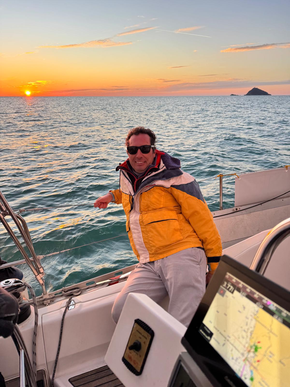 Uomo con giacca da vela gialla e occhiali scuri seduto nel pozzetto di una barca al tramonto, isolotto sull'orizzonte